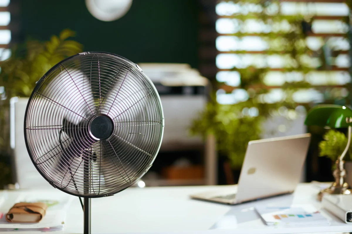 shutterstock_2441679465 1 | TVGURU.cz Large black desk fan on a white office desk with a laptop and papers in the background.