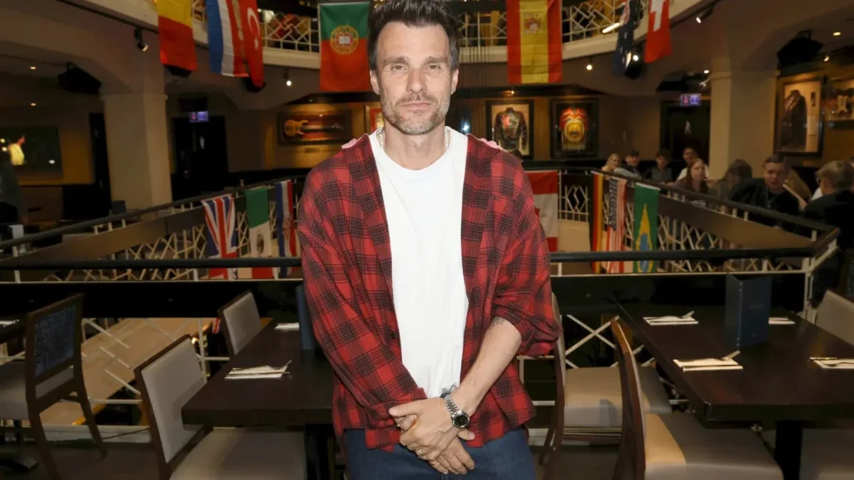 Man in a red plaid shirt over a white tee poses in a flag-filled event space or gallery.