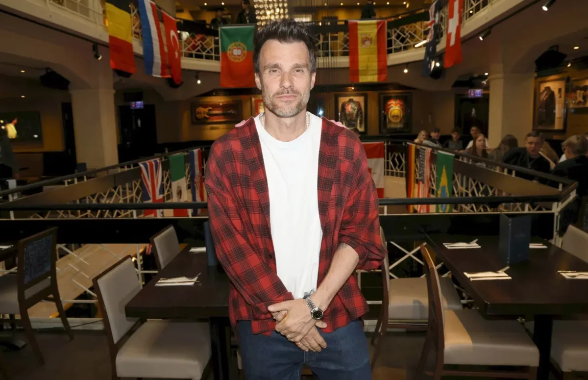 Man in a red plaid shirt over a white tee poses in a flag-filled event space or gallery.