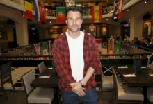 Man in a red plaid shirt over a white tee poses in a flag-filled event space or gallery.