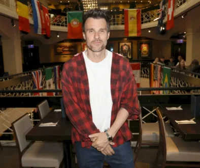 Man in a red plaid shirt over a white tee poses in a flag-filled event space or gallery.
