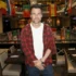 Man in a red plaid shirt over a white tee poses in a flag-filled event space or gallery.