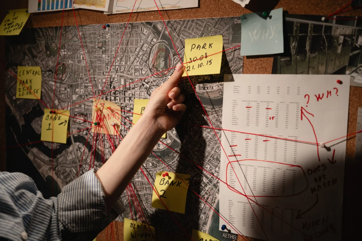 shutterstock_2794736381 1 | TVGURU.cz Hand reaching to place a yellow sticky note labeled'PARK' on a map-board connected by red strings and other notes (investigation board).