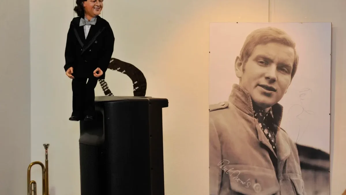 Ventriloquist dummy in a tuxedo perched on a speaker, beside a large black‑and‑white portrait of a man on the wall.