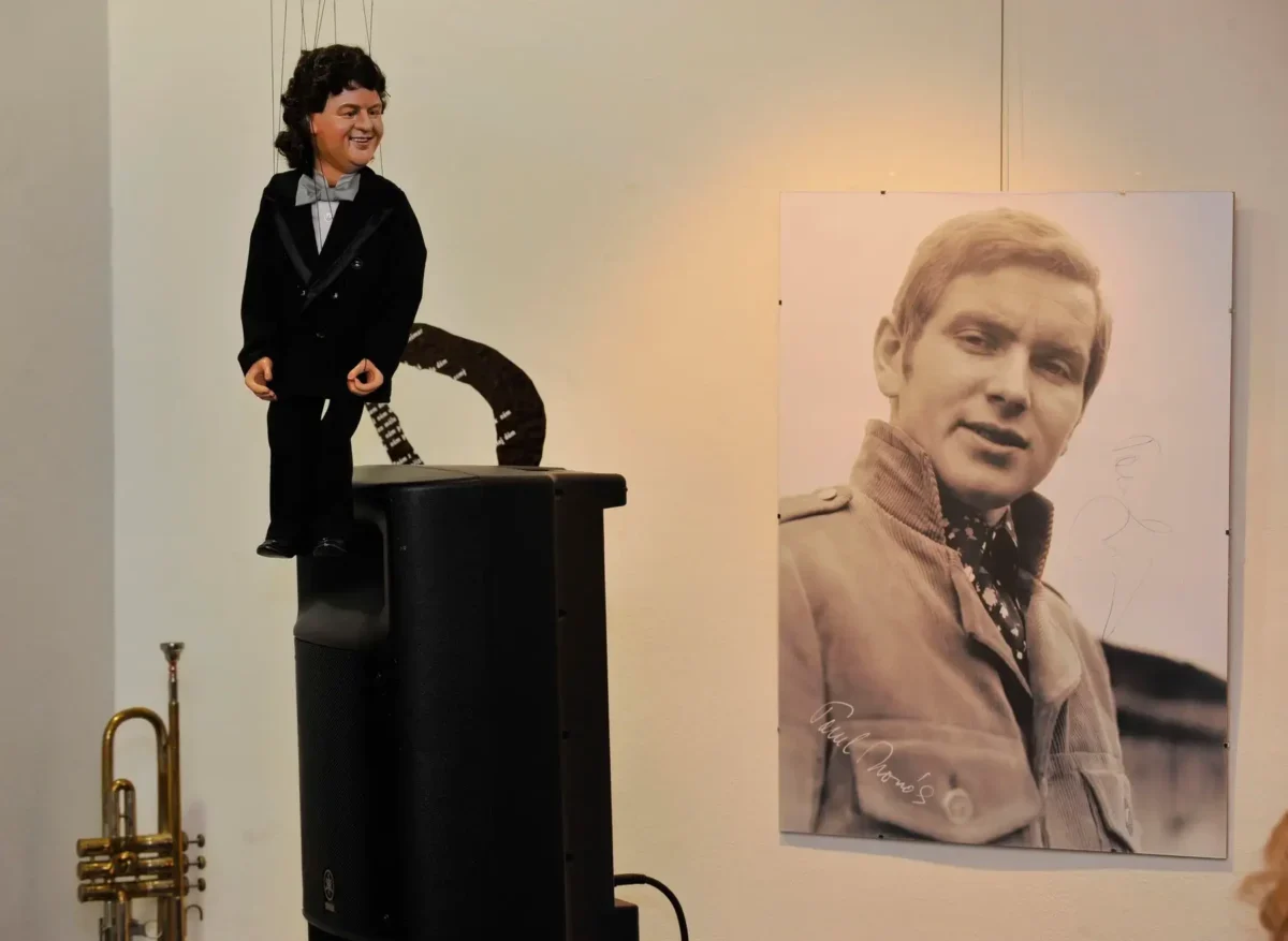 Ventriloquist dummy in a tuxedo perched on a speaker, beside a large black‑and‑white portrait of a man on the wall.