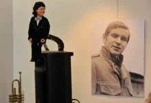 Ventriloquist dummy in a tuxedo perched on a speaker, beside a large black‑and‑white portrait of a man on the wall.