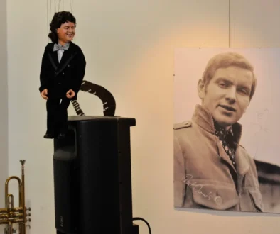 Ventriloquist dummy in a tuxedo perched on a speaker, beside a large black‑and‑white portrait of a man on the wall.