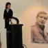 Ventriloquist dummy in a tuxedo perched on a speaker, beside a large black‑and‑white portrait of a man on the wall.