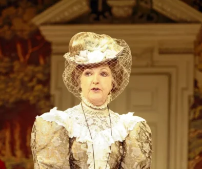 Woman in a vintage lace dress and a large hat with a net veil, performing on a stage with autumnal backdrop.