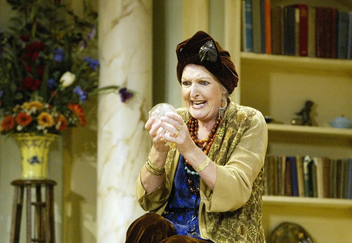 Older woman in elaborate outfit and brown turban, gazing at a crystal ball during a theatrical performance in a cozy setting with flowers and books nearby.