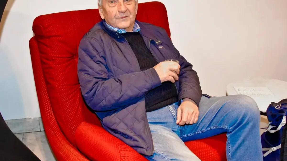 Older man with white hair sits in a red armchair, wearing a blue quilted jacket and jeans, holding a glass in his hand.