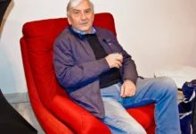 Older man with white hair sits in a red armchair, wearing a blue quilted jacket and jeans, holding a glass in his hand.