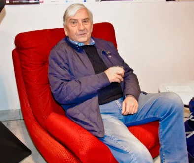 Older man with white hair sits in a red armchair, wearing a blue quilted jacket and jeans, holding a glass in his hand.