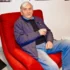 Older man with white hair sits in a red armchair, wearing a blue quilted jacket and jeans, holding a glass in his hand.