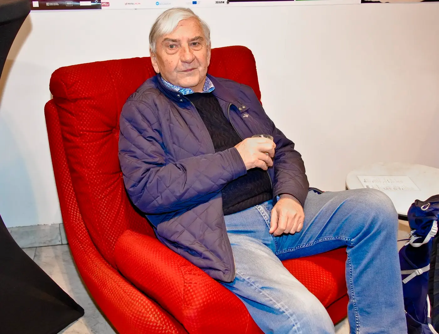 Older man with white hair sits in a red armchair, wearing a blue quilted jacket and jeans, holding a glass in his hand.