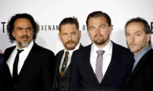Four men in black suits posing together at a formal event/photo backdrop on a red carpet area.