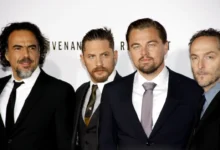 Four men in black suits posing together at a formal event/photo backdrop on a red carpet area.