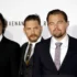 Four men in black suits posing together at a formal event/photo backdrop on a red carpet area.