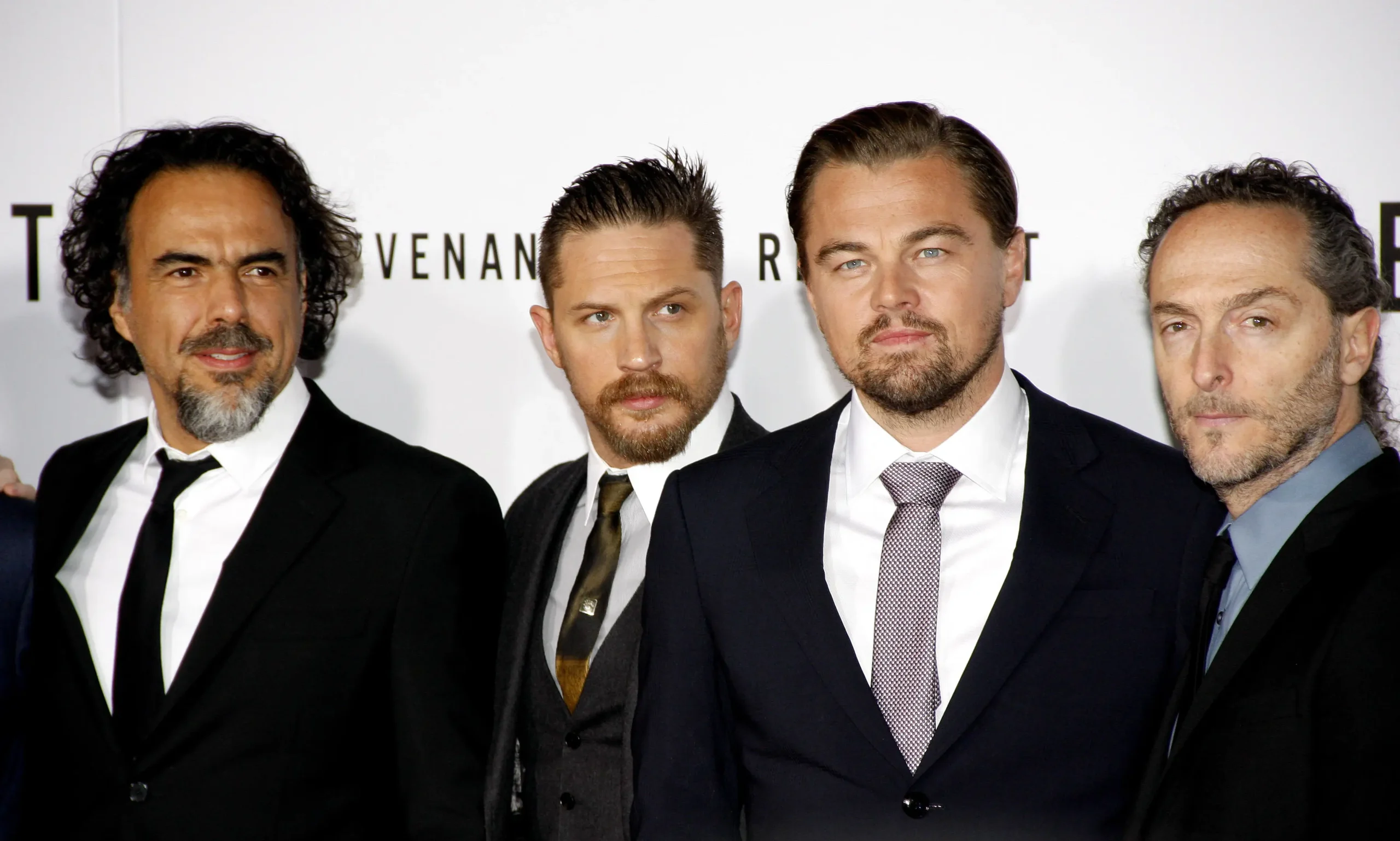 Four men in black suits posing together at a formal event/photo backdrop on a red carpet area.