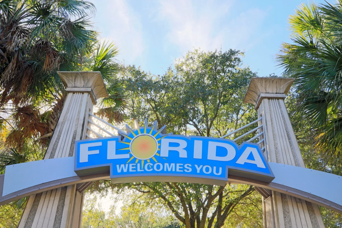 shutterstock_2258377307 1 | TVGURU.cz Entrance sign reading'Florida welcomes you' with a sun emblem, framed by palm trees and concrete columns under a blue sky.