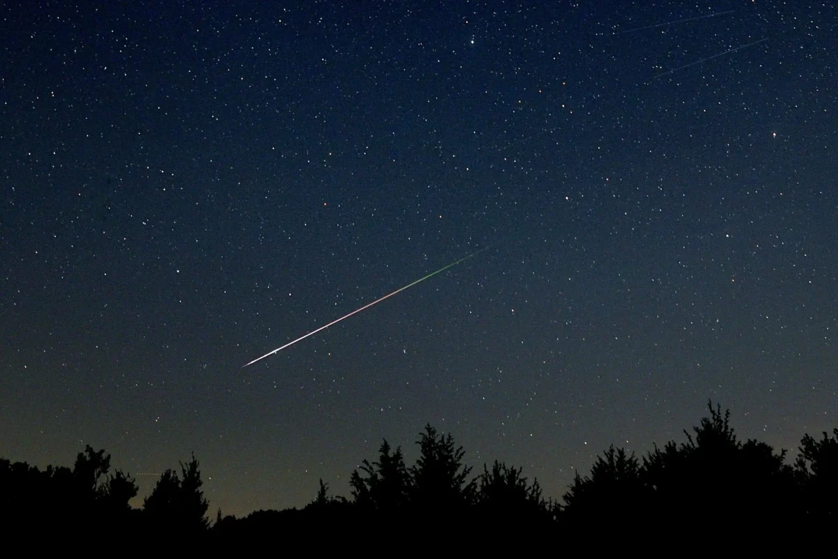 Starry night sky with a bright meteor streak crossing diagonally above a dark forest silhouette.