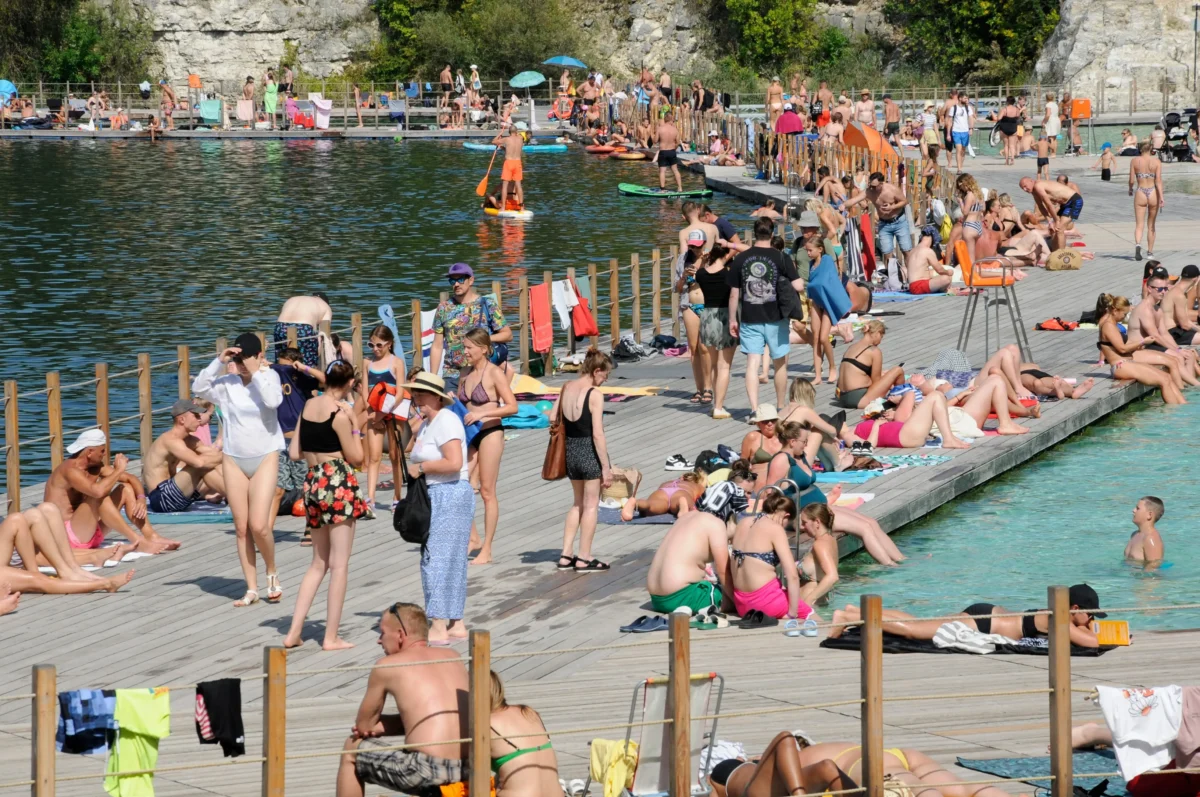 Crowded lakeside boardwalk with sunbathers on towels, swimmers in water, and people using inflatable paddles on a sunny day by rocky cliffs.