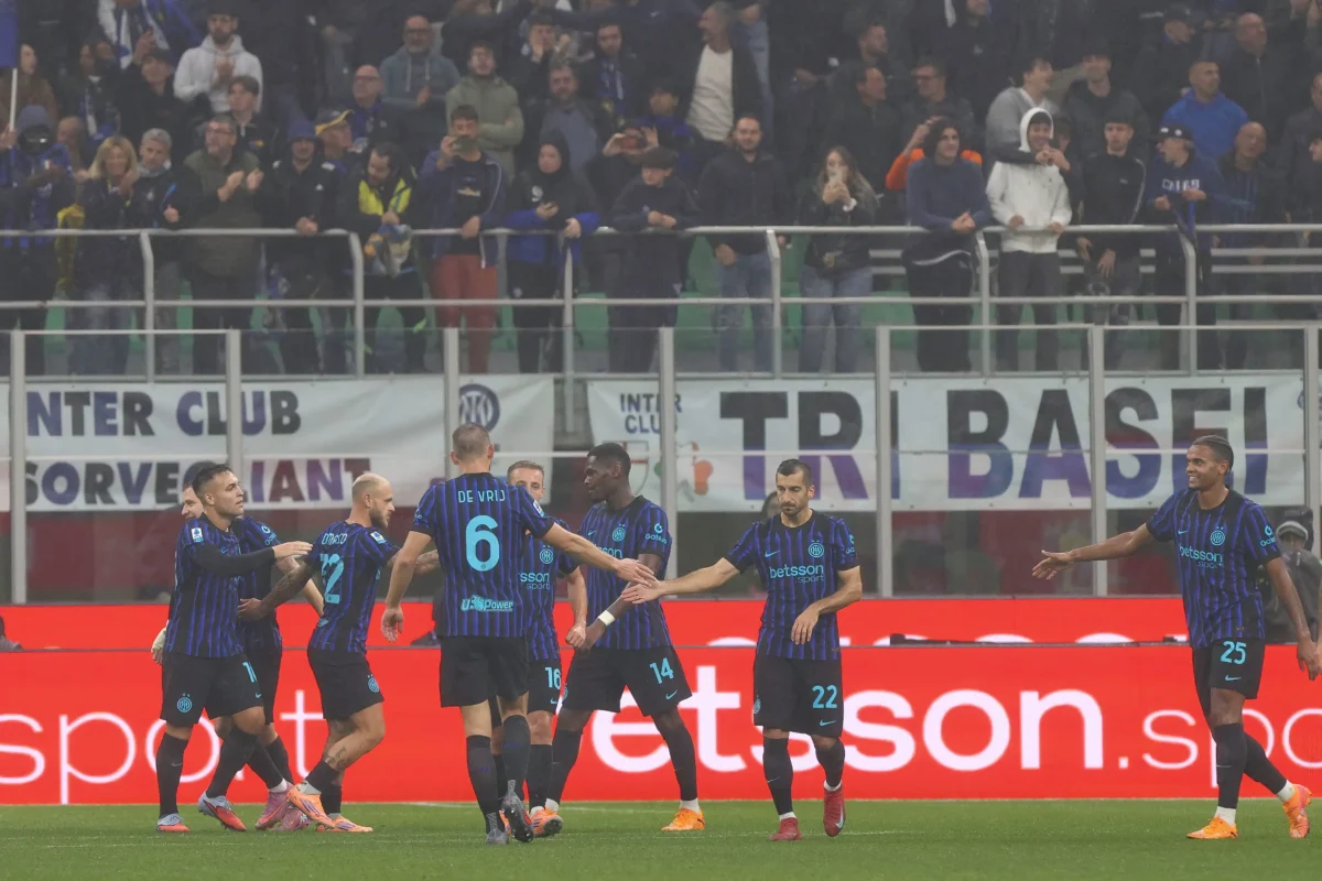 shutterstock_2686880929 1 | TVGURU.cz Soccer players in blue-black striped kits celebrate on the pitch with fans cheering in the stands behind them.