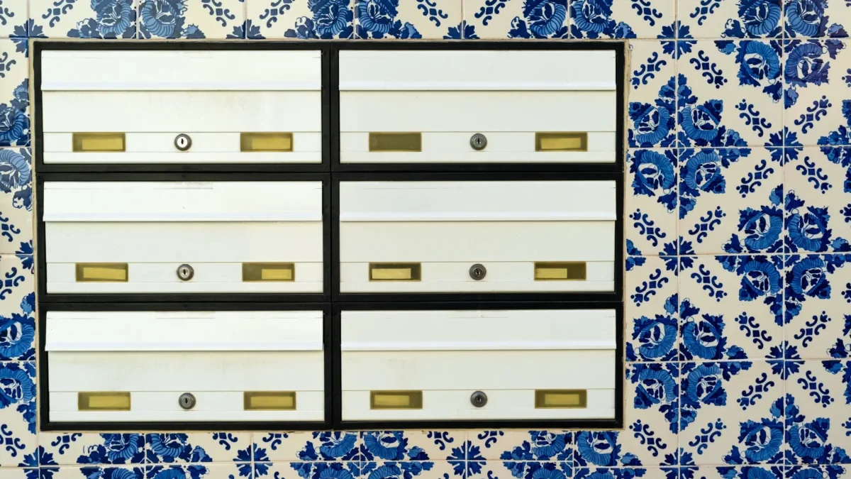 Row of white metal mailboxes with brass slots and locks, mounted on a black frame against a blue-and-white tiled wall.