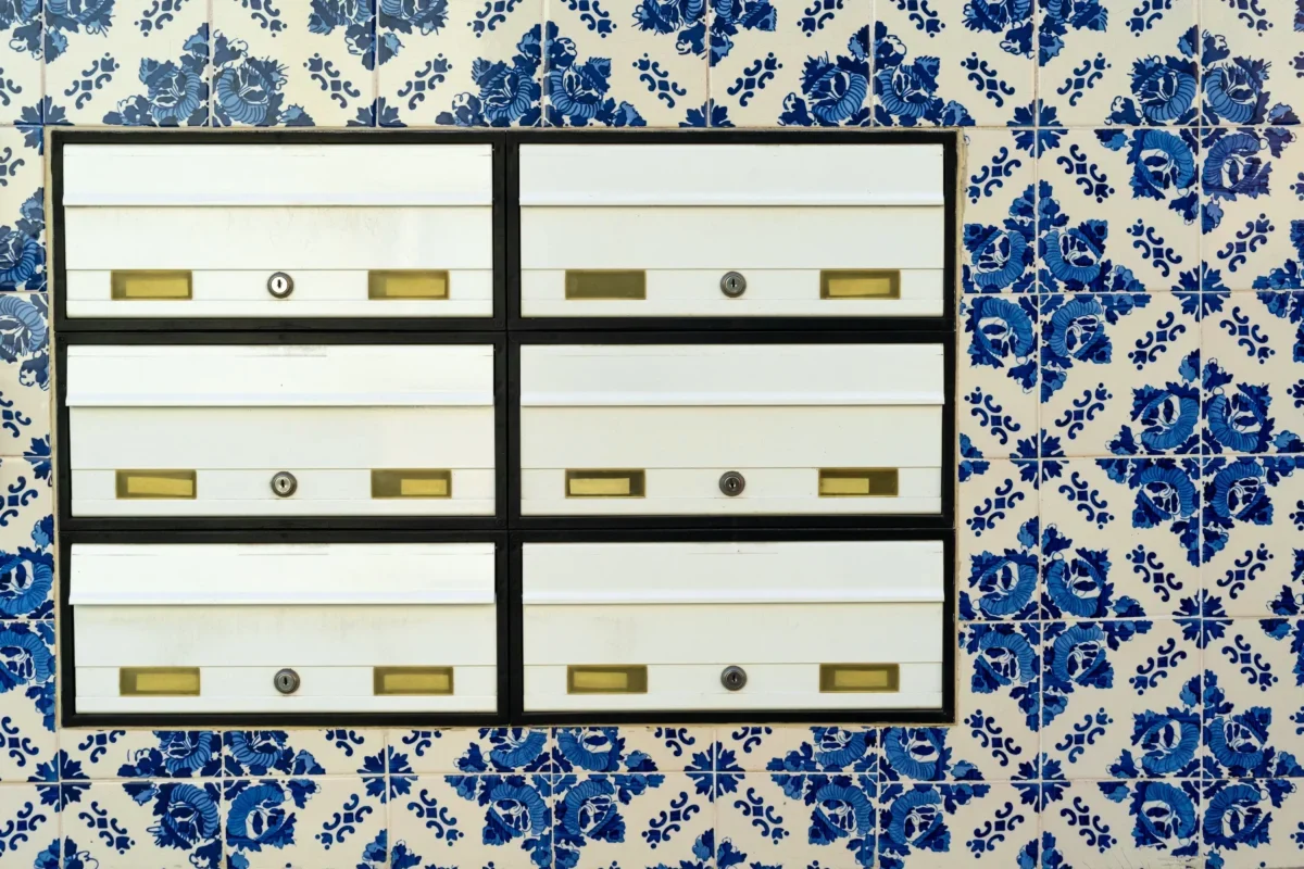Row of white metal mailboxes with brass slots and locks, mounted on a black frame against a blue-and-white tiled wall.