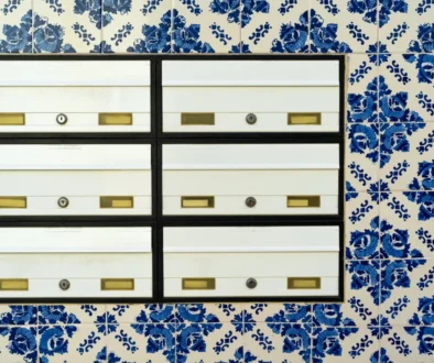 Row of white metal mailboxes with brass slots and locks, mounted on a black frame against a blue-and-white tiled wall.