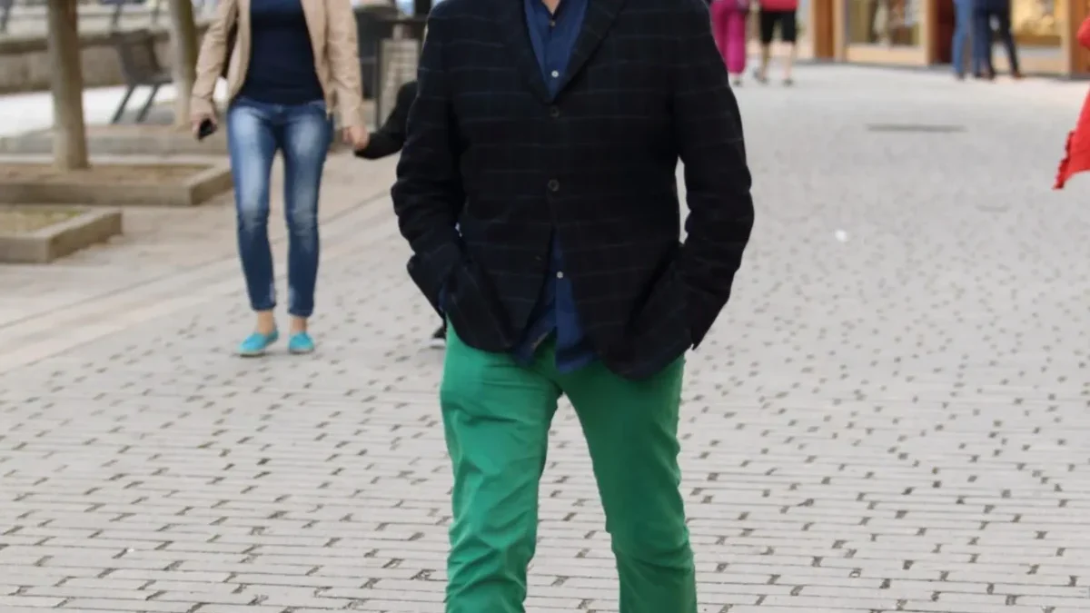 Man in a dark plaid blazer and bright green pants walks toward the camera on a cobblestone street, with a woman in a beige coat behind him.