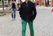 Man in a dark plaid blazer and bright green pants walks toward the camera on a cobblestone street, with a woman in a beige coat behind him.