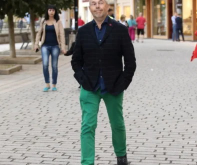 Man in a dark plaid blazer and bright green pants walks toward the camera on a cobblestone street, with a woman in a beige coat behind him.