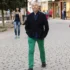 Man in a dark plaid blazer and bright green pants walks toward the camera on a cobblestone street, with a woman in a beige coat behind him.