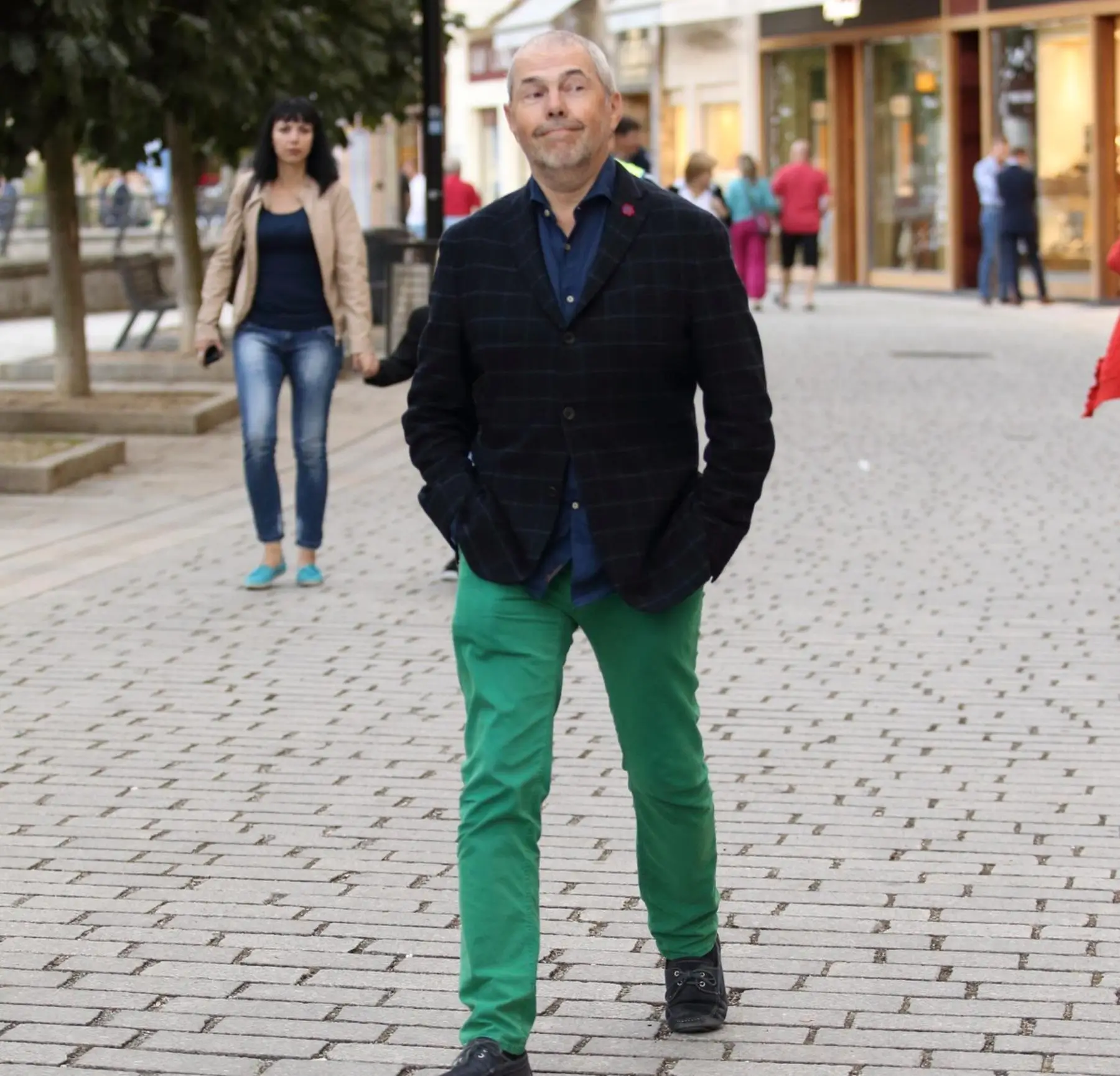 Man in a dark plaid blazer and bright green pants walks toward the camera on a cobblestone street, with a woman in a beige coat behind him.