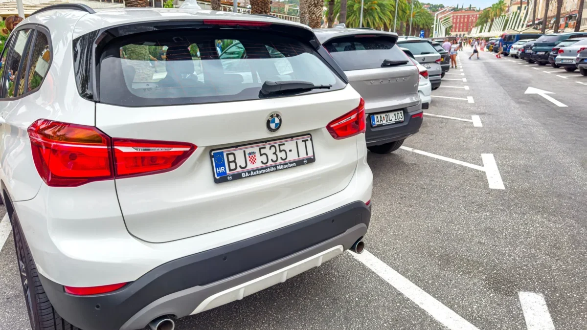 Rear view of a white BMW SUV parked in a crowded street; palm trees line the sidewalk and pedestrians walk in the distance.