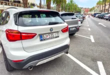 Rear view of a white BMW SUV parked in a crowded street; palm trees line the sidewalk and pedestrians walk in the distance.
