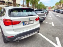 Rear view of a white BMW SUV parked in a crowded street; palm trees line the sidewalk and pedestrians walk in the distance.