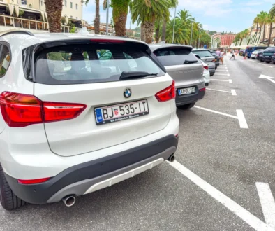 Rear view of a white BMW SUV parked in a crowded street; palm trees line the sidewalk and pedestrians walk in the distance.