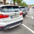 Rear view of a white BMW SUV parked in a crowded street; palm trees line the sidewalk and pedestrians walk in the distance.