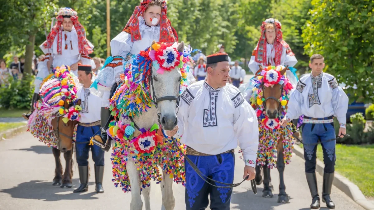 Group of people in traditional embroidered costumes riding horses decorated with rainbow paper garlands in a festive parade outdoors.