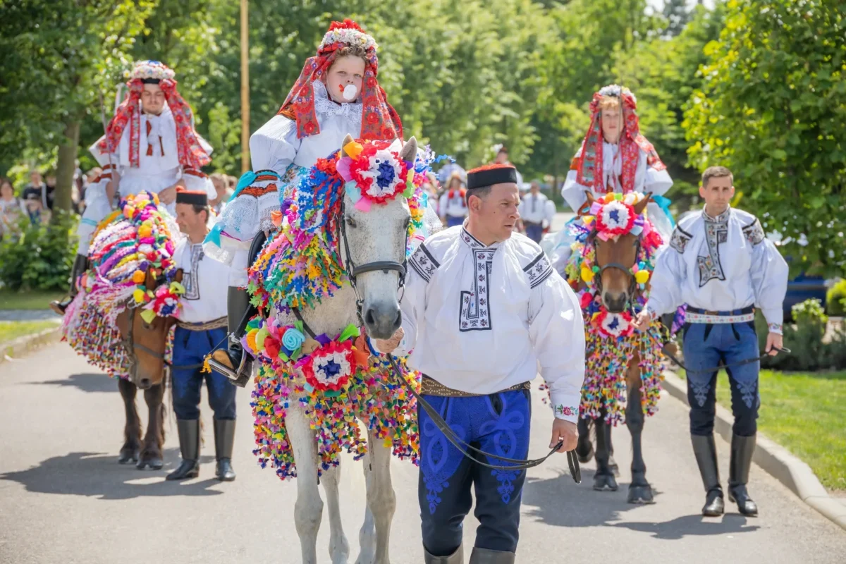 Group of people in traditional embroidered costumes riding horses decorated with rainbow paper garlands in a festive parade outdoors.