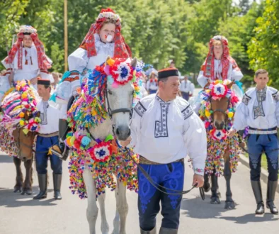 Group of people in traditional embroidered costumes riding horses decorated with rainbow paper garlands in a festive parade outdoors.