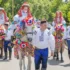 Group of people in traditional embroidered costumes riding horses decorated with rainbow paper garlands in a festive parade outdoors.