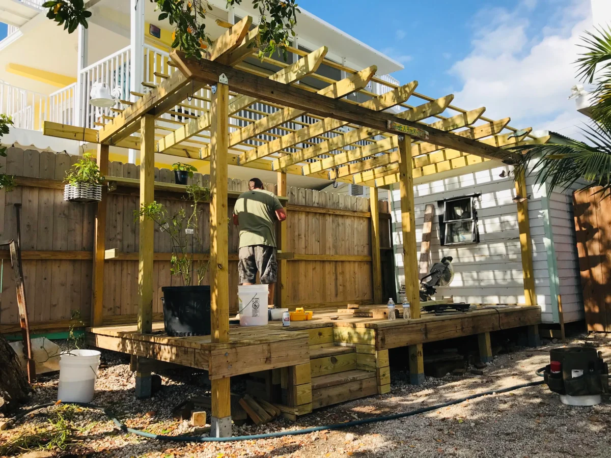 Backyard construction scene with a wooden pergola frame over a raised deck; a man in a green shirt works on the structure with buckets nearby.
