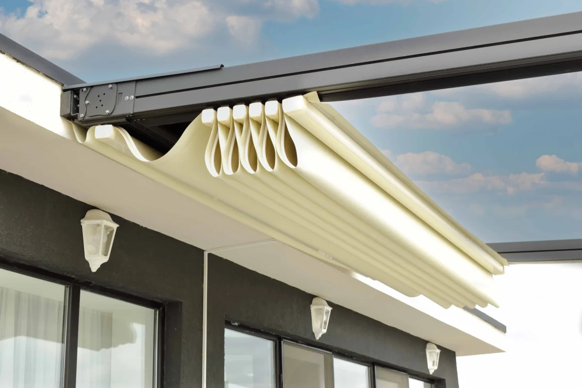 Close-up of a white retractable awning with folded fabric panels above glass doors on a dark exterior wall, under a blue sky.