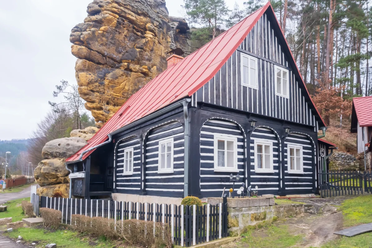 Two-story black-and-white striped house with a bright red metal roof beside a large rock cliff and trees in the background, with a wooden fence in front.