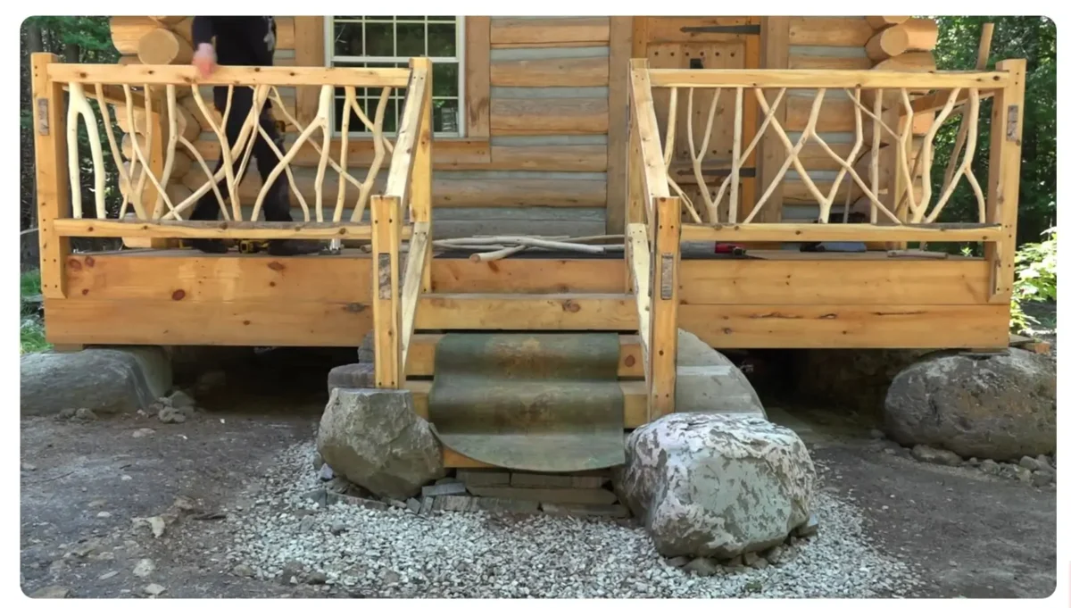 Bez názvu 1 | TVGURU.cz Rustic wooden cabin porch with curved branch-like railings and stone steps leading up from a rocky ground surface