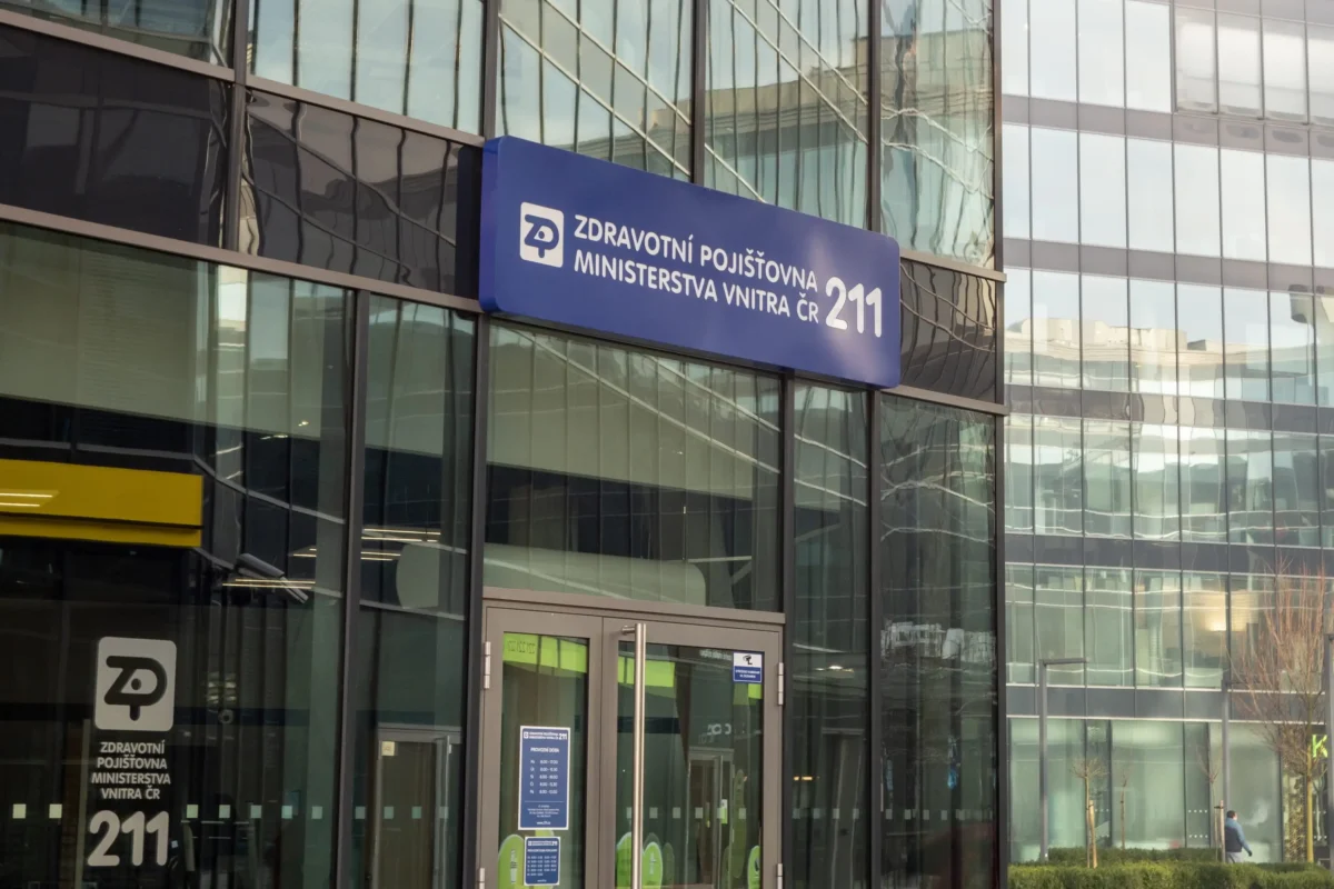 shutterstock_1618442716 1 | TVGURU.cz Blue sign on a glass office building reading'Zdravotní pojišťovna Ministerstva vnitra ČR 211' with logo nearby.
