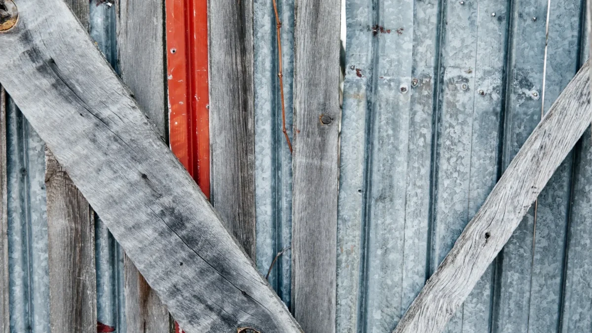 Weathered vertical wooden slats with peeling gray paint, crossed by a diagonal weathered board and a narrow red strip visible behind.